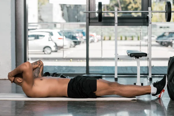 Hombre afroamericano tatuado haciendo abdominales en la colchoneta de fitness en el gimnasio - foto de stock