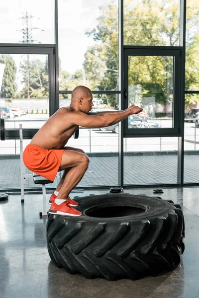 Side view of african american athlete doing squats on tire at gym — Stock Photo