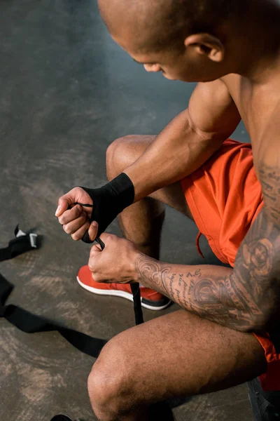 Visão de alto ângulo de tatuado esportista afro-americano envolvendo mão em bandagem de boxe no ginásio — Fotografia de Stock