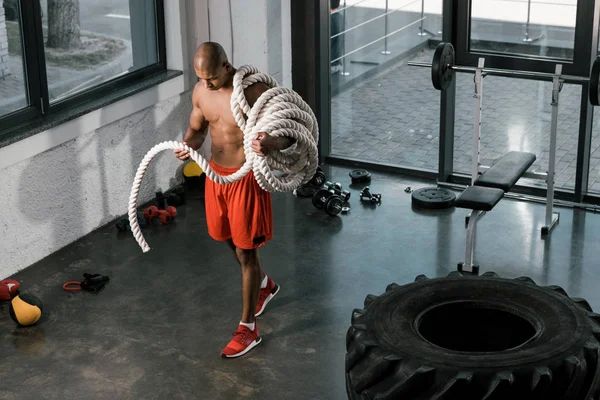 Vista de ángulo alto del atleta afroamericano muscular sin camisa con cuerda de batalla en el gimnasio - foto de stock