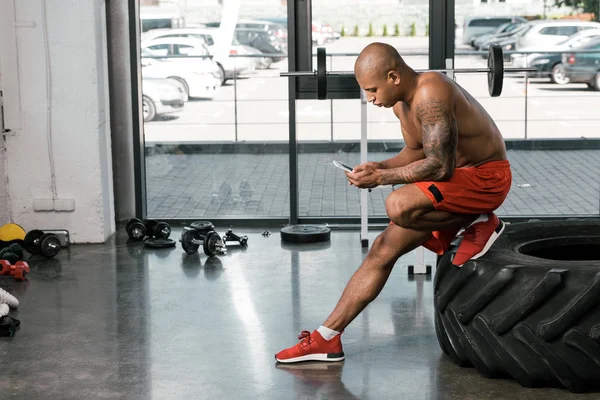 Vista lateral do atleta afro-americano sem camisa descansando e usando smartphone no pneu no ginásio — Fotografia de Stock