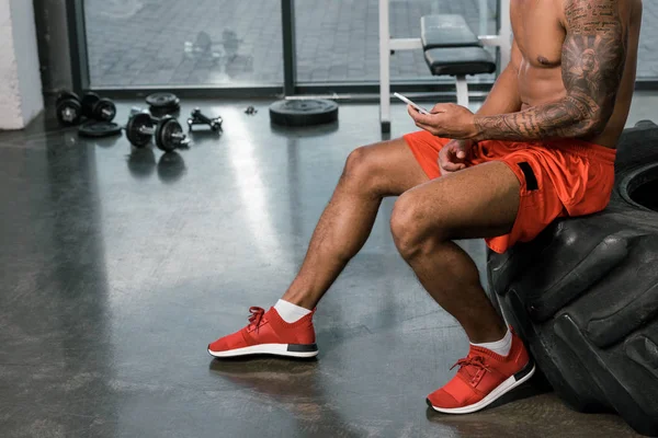 Imagen recortada de atleta afroamericano tatuado descansando y usando teléfono inteligente en el neumático en el gimnasio - foto de stock