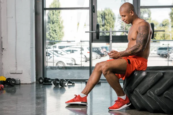 Vue latérale de l'homme afro-américain heureux reposant et utilisant smartphone sur pneu à la salle de gym — Photo de stock