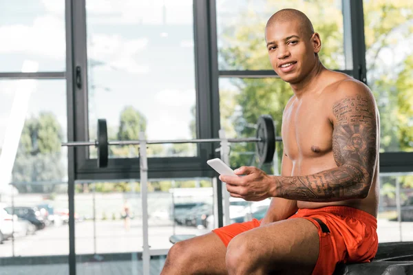 Sportif afro-américain souriant avec la main tatouée tenant smartphone et regardant la caméra dans la salle de gym — Photo de stock