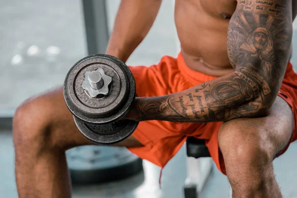 Imagen recortada de atleta afroamericano tatuado haciendo ejercicio con mancuerna en el gimnasio - foto de stock
