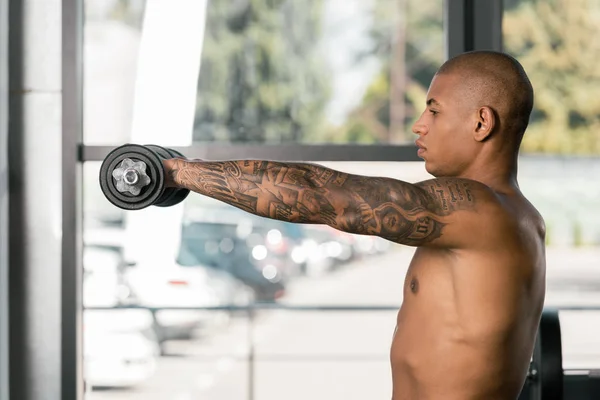 Selective focus of muscular shirtless african american sportsman exercising with dumbbells at gym — Stock Photo