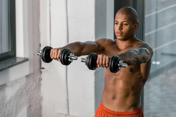 Молодий м'язистий без сорочки афроамериканський спортсмен займається з гантелями в спортзалі — стокове фото