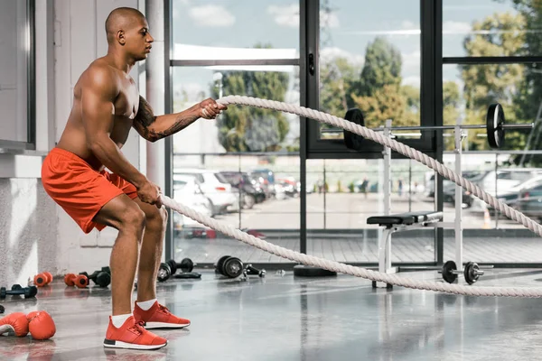 Vista lateral del atleta afroamericano haciendo ejercicio con cuerdas de batalla en el gimnasio - foto de stock