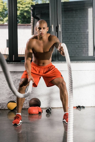 Musculoso sin camisa atleta afroamericano haciendo ejercicio con cuerdas de batalla en el gimnasio - foto de stock