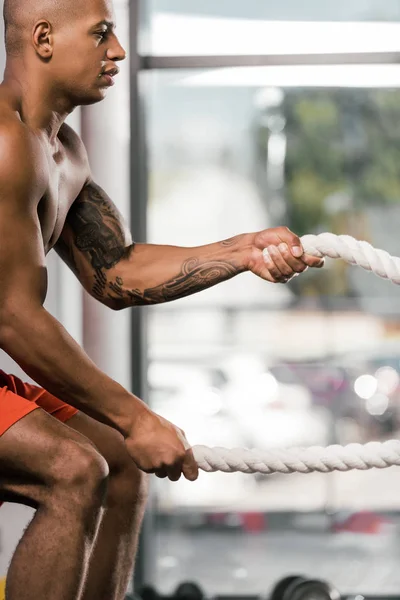 Vista lateral del atleta afroamericano haciendo ejercicio con cuerdas de batalla en el gimnasio - foto de stock