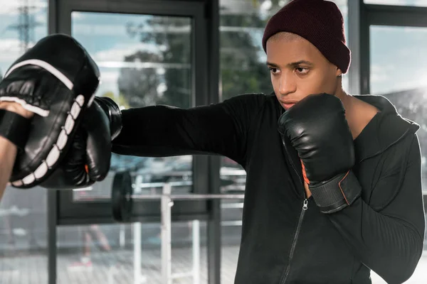 Молодой африканский американский боксер в боксёрских перчатках тренируется с тренером в тренажерном зале — стоковое фото