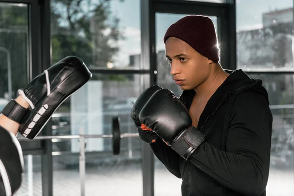 Sério boxeador afro-americano em luvas de boxe praticando com treinador no ginásio — Fotografia de Stock