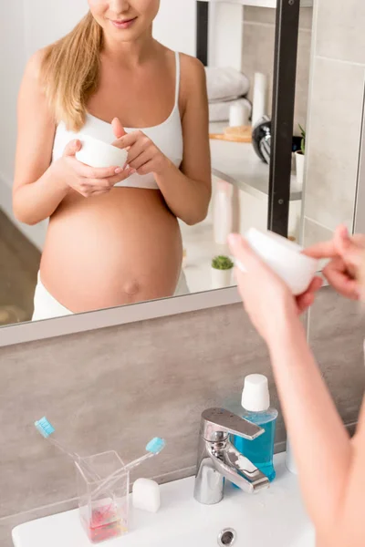 Recortado disparo de sonriente mujer embarazada con tarro de crema de pie delante del espejo en el baño - foto de stock