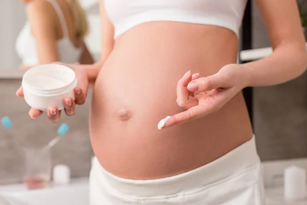 Plan recadré de femme enceinte avec crème près du ventre pour éviter les vergetures dans la salle de bain — Photo de stock