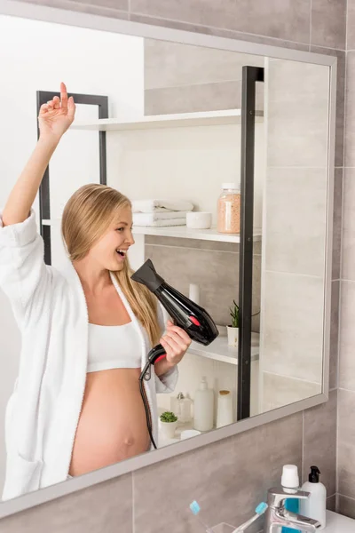 Happy beautiful pregnant woman in bathrobe singing at hair dryer in front of mirror in bathroom — Stock Photo