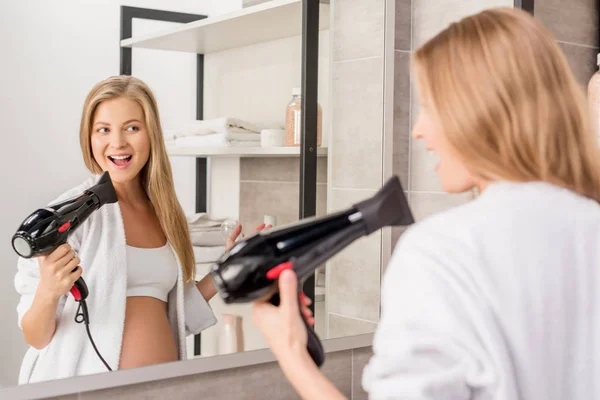 Happy pregnant woman in bathrobe singing at hair dryer in front of mirror in bathroom — Stock Photo