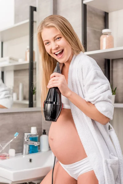 Happy pregnant woman singing at hair dryer in bathroom — Stock Photo
