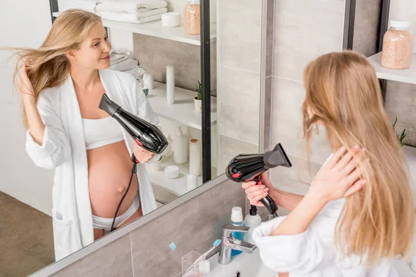 Vue grand angle de belle femme enceinte séchant les cheveux tout en regardant miroir dans la salle de bain — Photo de stock
