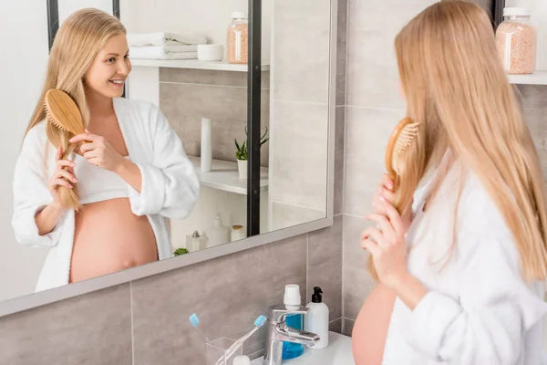 Felice donna incinta in accappatoio spazzolarsi i capelli e guardando specchio in bagno — Foto stock