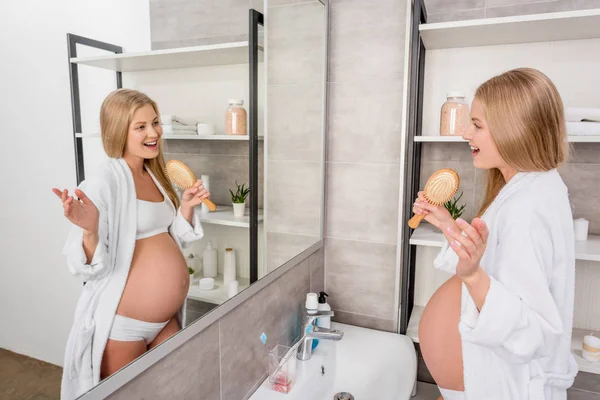 Счастливая беременная женщина в нижнем белье поет в расческе и смотрит в зеркало в ванной комнате — стоковое фото