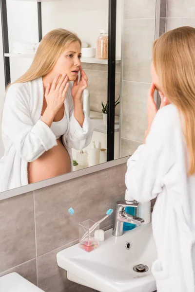 Depressed pregnant woman with skin problems looking at her face through mirror in bathroom — Stock Photo