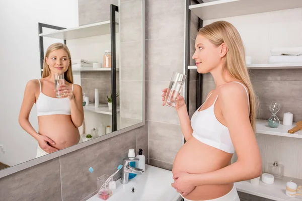Щаслива вагітна жінка зі склянкою води дивиться на дзеркало у ванній — стокове фото