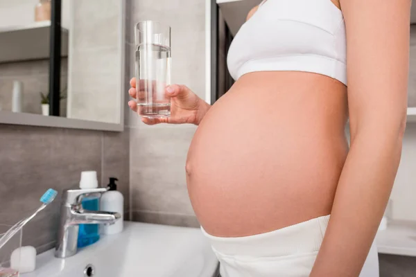 Обрезанный снимок беременной женщины в нижнем белье со стаканом воды, стоящей в ванной комнате — стоковое фото