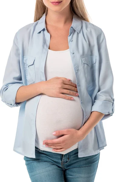 Обрезанный снимок улыбающейся беременной женщины в джинсах и рубашке, держащей животик с изолированными на белом руками — стоковое фото