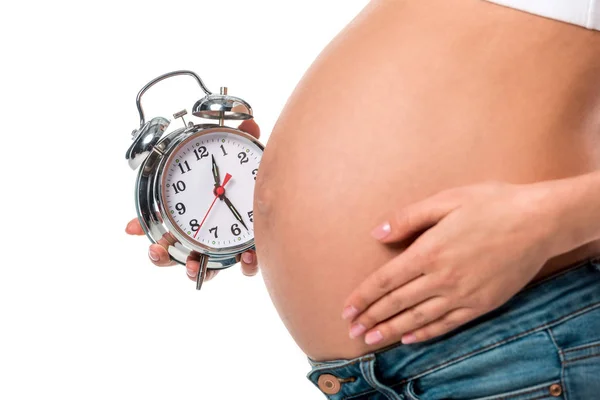 Vista parcial de la mujer embarazada sosteniendo el despertador cerca del vientre aislado en blanco - foto de stock