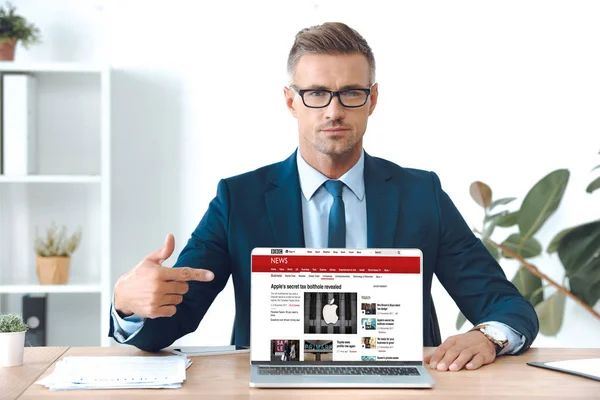 Guapo hombre de negocios en gafas apuntando a la computadora portátil con el sitio web de noticias de la penes en la pantalla - foto de stock