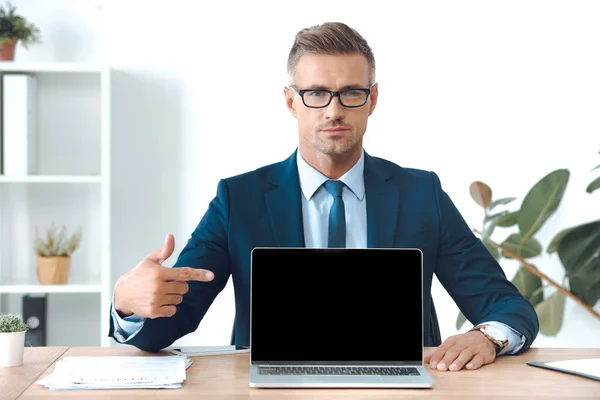 Hombre de negocios apuntando a la computadora portátil con pantalla en blanco y mirando a la cámara - foto de stock