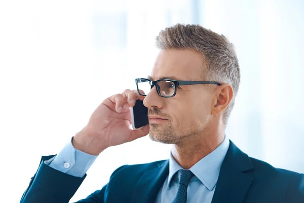 Apuesto hombre de negocios confiado en gafas hablando por teléfono inteligente y mirando hacia otro lado en la oficina - foto de stock