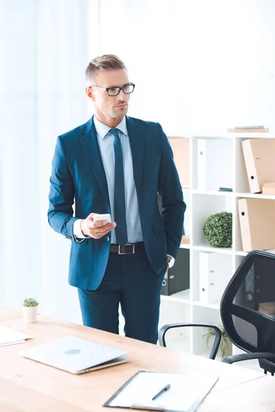 Uomo d'affari fiducioso in occhiali tenendo smartphone e guardando lontano in ufficio — Foto stock