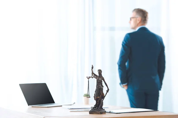 Portátil con pantalla en blanco y dama estatua de la justicia en la mesa y el abogado mirando a la ventana detrás - foto de stock