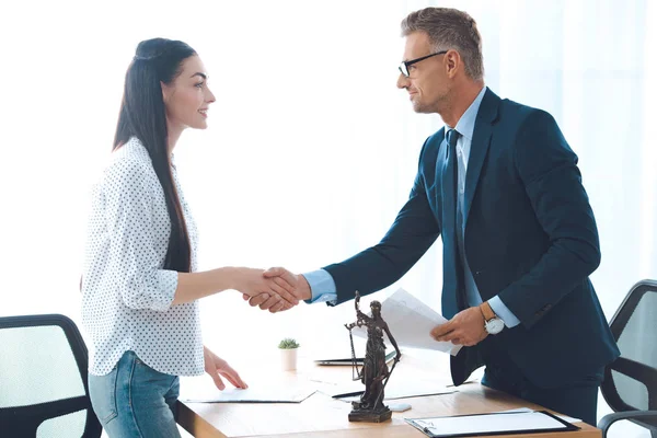 Vista lateral de abogado profesional y cliente joven mujer estrechando la mano en la oficina - foto de stock