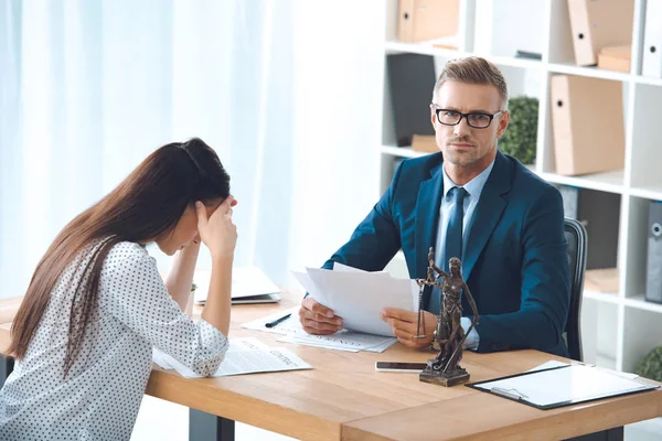 Abogado sosteniendo papeles y mirando a la cámara mientras triste cliente femenino sentado en la oficina - foto de stock