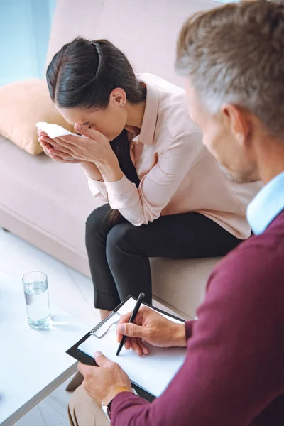 Vue grand angle de psychologue écrivant sur presse-papiers et regardant patient pleurer sur le canapé — Photo de stock