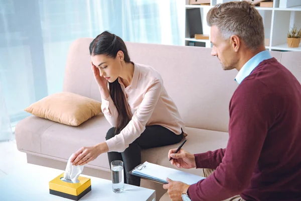 Psychotherapist taking notes on clipboard while patient sitting on couch and crying — Stock Photo