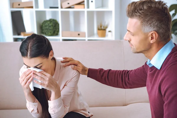 Psicoterapeuta que apoya a un paciente joven sentado en el sofá y llorando - foto de stock