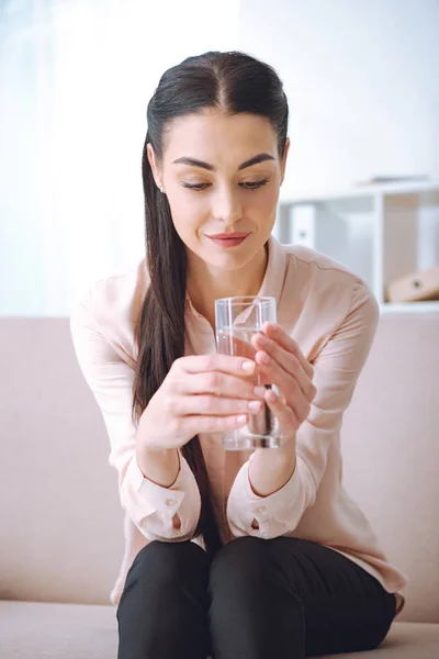 Attraktive lächelnde junge Frau sitzt auf der Couch und hält ein Glas Wasser in der Hand — Stockfoto