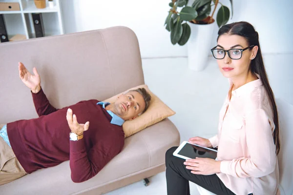 Психотерапевт в очках с помощью цифрового планшета и глядя на камеру, пока пациент лежит на диване — стоковое фото