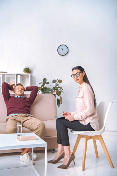 Atractivo psicoterapeuta mirando a la cámara mientras el paciente descansa con las manos detrás de la cabeza en el sofá - foto de stock