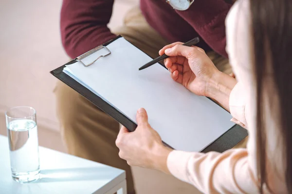 Partial view of female psychologist pointing at blank clipboard during therapy appointment with male patient — Stock Photo