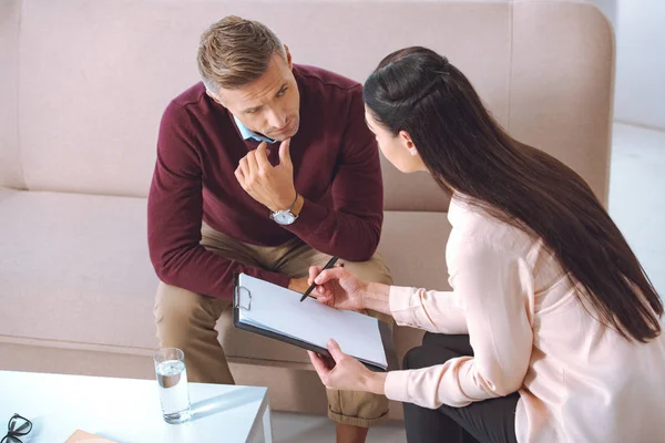 Psychologue femelle pointant vers le presse-papiers vierge pendant le rendez-vous de thérapie avec le patient masculin — Photo de stock