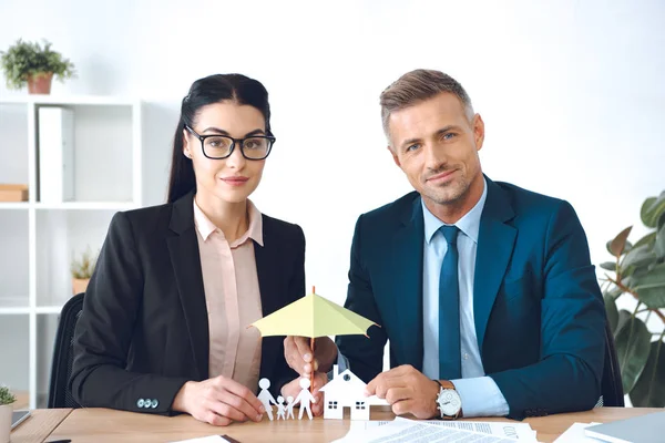 Insurance agents covering family and house paper models with umbrella at workplace — Stock Photo