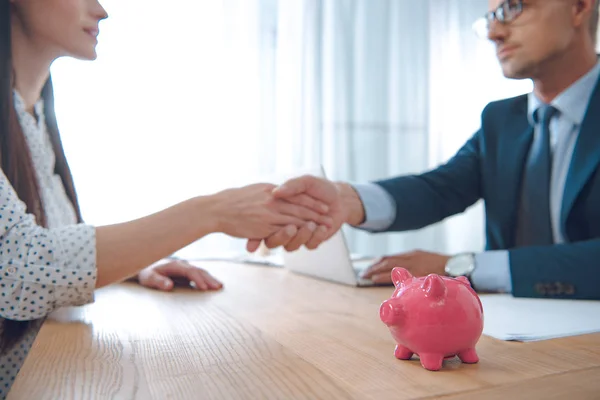 Foco seletivo do agente de seguros e do cliente apertando as mãos na mesa com o banco piggy rosa — Fotografia de Stock