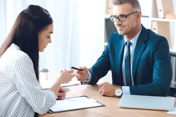 Smiling insurance agent giving pen to female client for signing papers at tabletop in office — Stock Photo