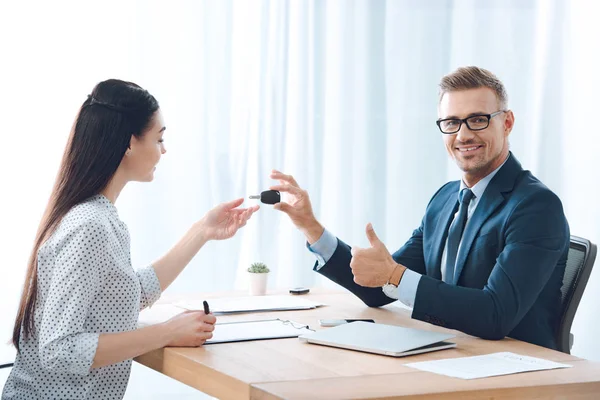 Agente de seguros sorrindo dando chave de carro para cliente feminino no local de trabalho no escritório — Fotografia de Stock