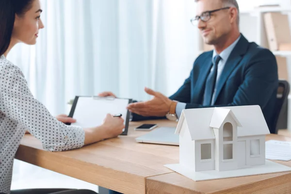 Agente de seguros en anteojos y cliente femenino que se reúne en el lugar de trabajo con el modelo de casa de papel en la oficina, concepto de seguro de casa - foto de stock