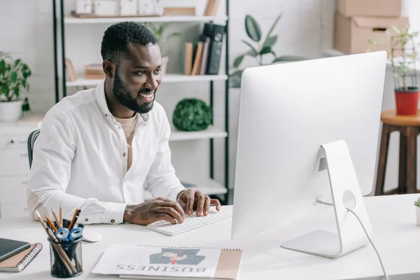 Улыбающийся красивый африканский бизнесмен, работающий за компьютером в офисе — стоковое фото